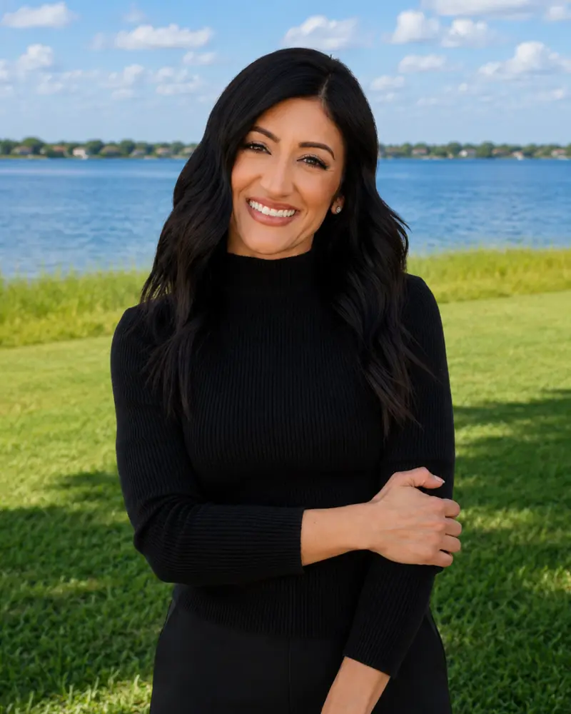 Smiling woman with long dark hair in a black top standing on a grassy shoreline by a blue lake, with trees and a partly cloudy sky in the background.