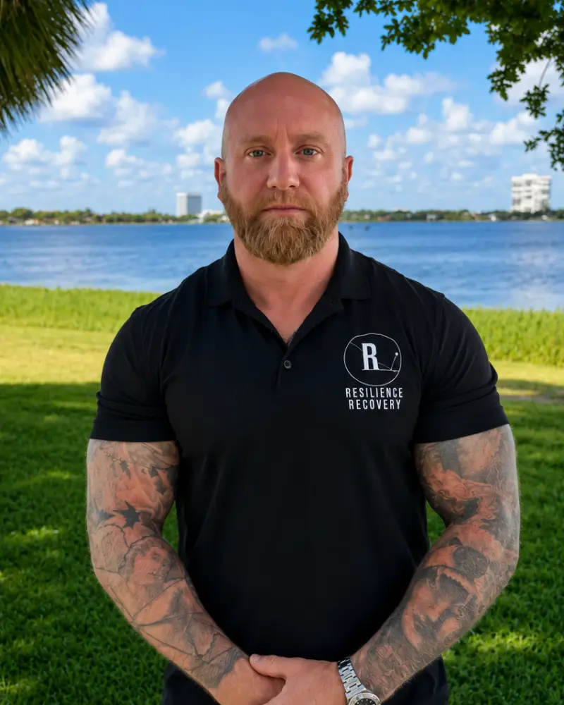 A bald, bearded man with tattooed arms standing outdoors on a grassy lawn facing the camera, wearing a black polo shirt with a “Resilience Recovery” logo by a large body of water under a blue sky.