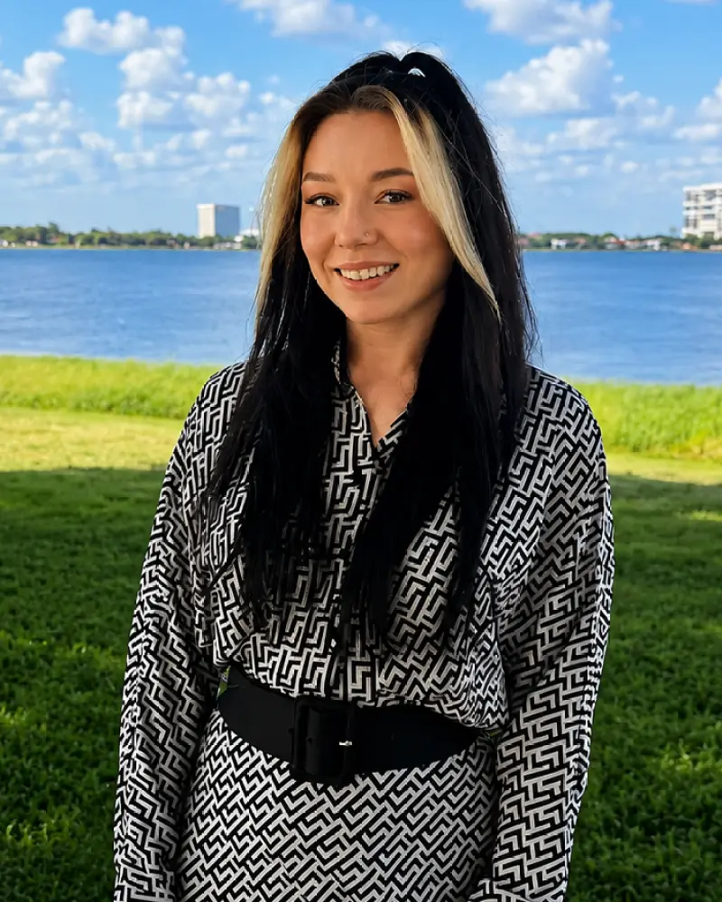 Smiling woman with long black-and-blonde hair standing outdoors near a grassy shoreline with a blue lake and buildings in the distance.
