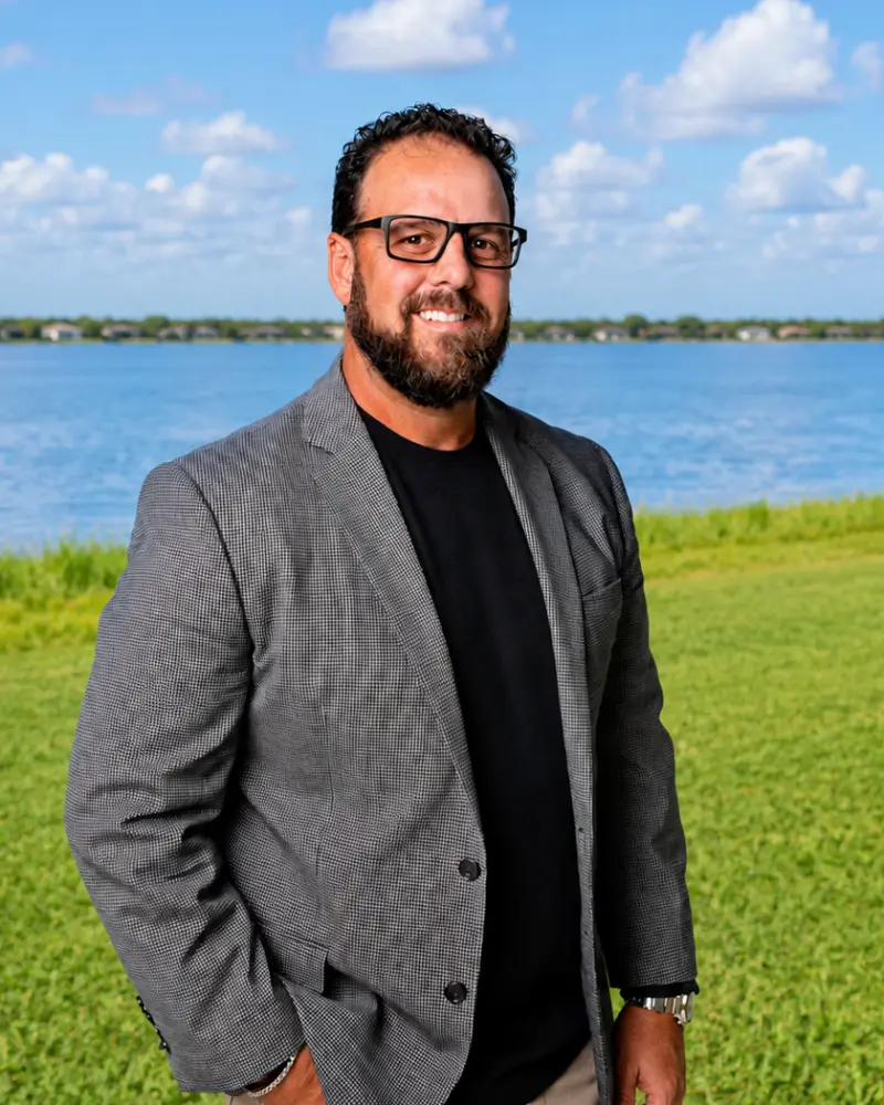 A smiling man with dark hair and glasses standing outdoors near a blue lake, wearing a gray blazer over a black shirt.