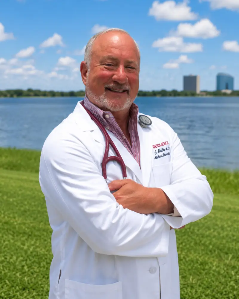 A smiling older male doctor in a white lab coat with a stethoscope stands with arms crossed outdoors by a lake, with a city skyline and blue sky in the background.