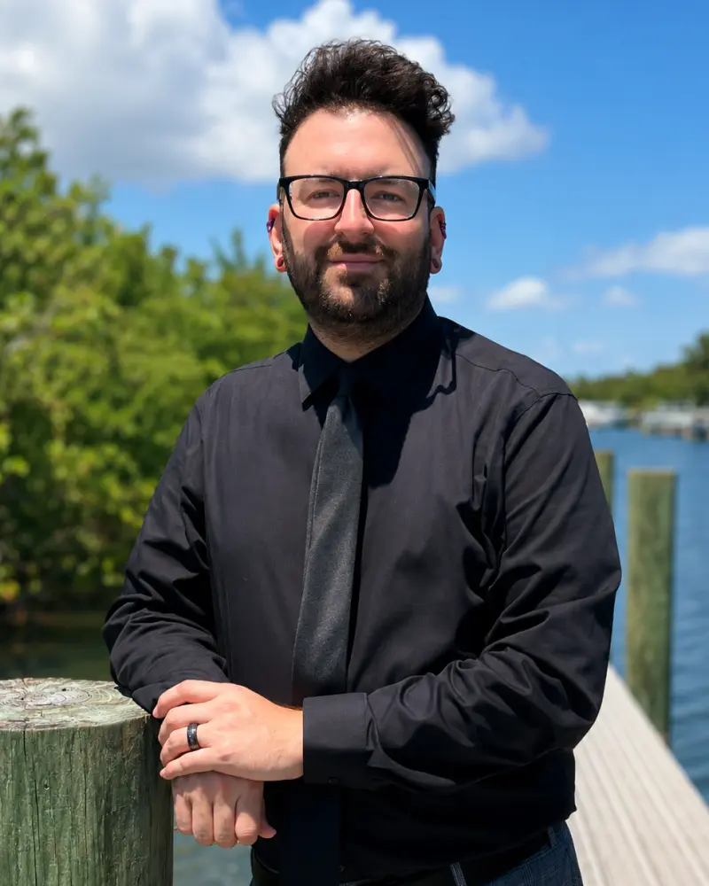A man with short dark hair and a beard wearing black glasses and a dark shirt standing outdoors by a wooden railing near a lake.