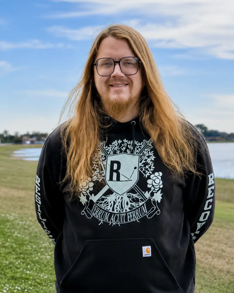 A man with long blonde hair and a beard wearing black glasses and a black hoodie with a crest logo, standing outdoors on a grassy field near water under a cloudy sky.