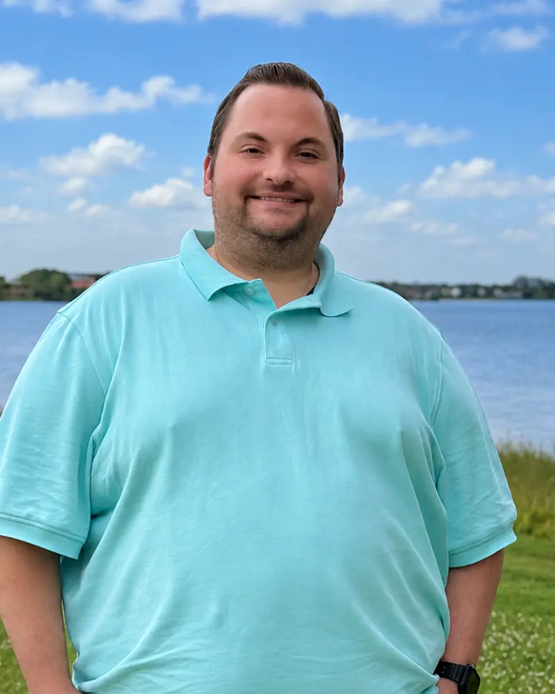 A smiling man in a turquoise polo shirt standing outdoors near a lake under a blue sky with scattered clouds.