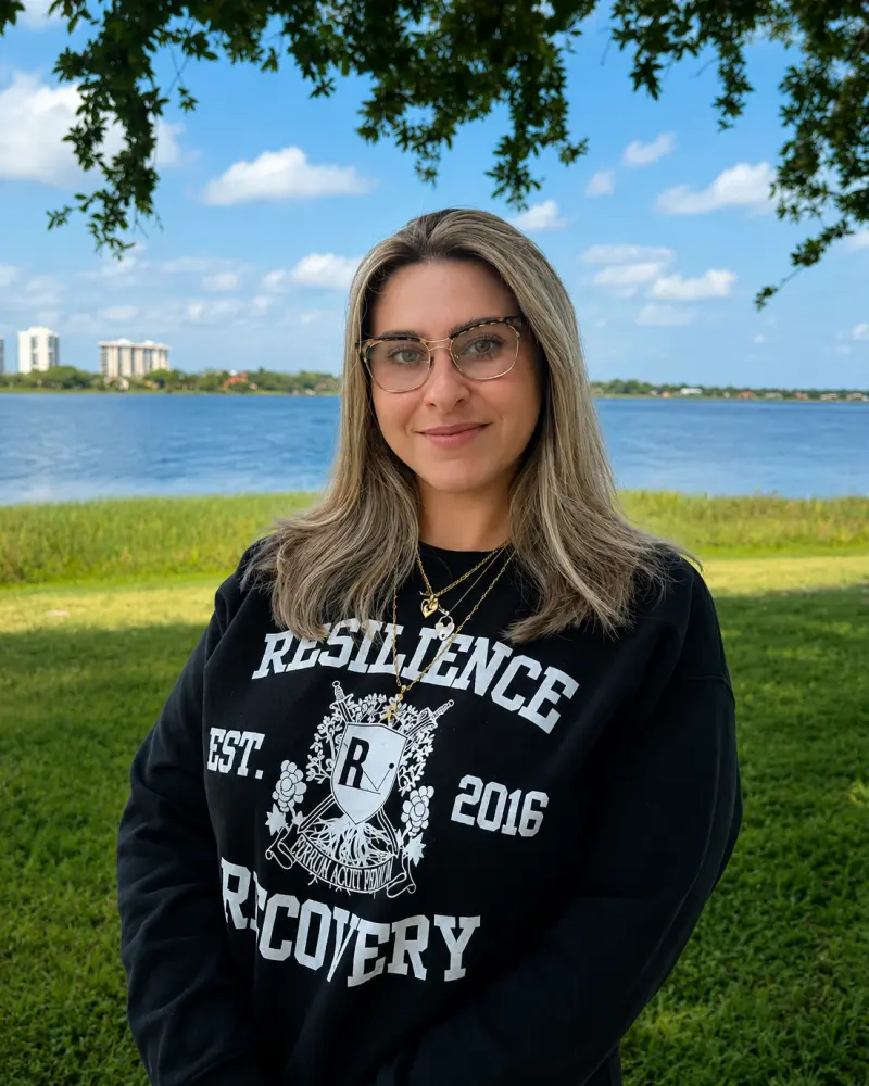 A woman wearing glasses and a black sweatshirt stands outdoors by a lake, with green grass and buildings in the distance under a blue sky.