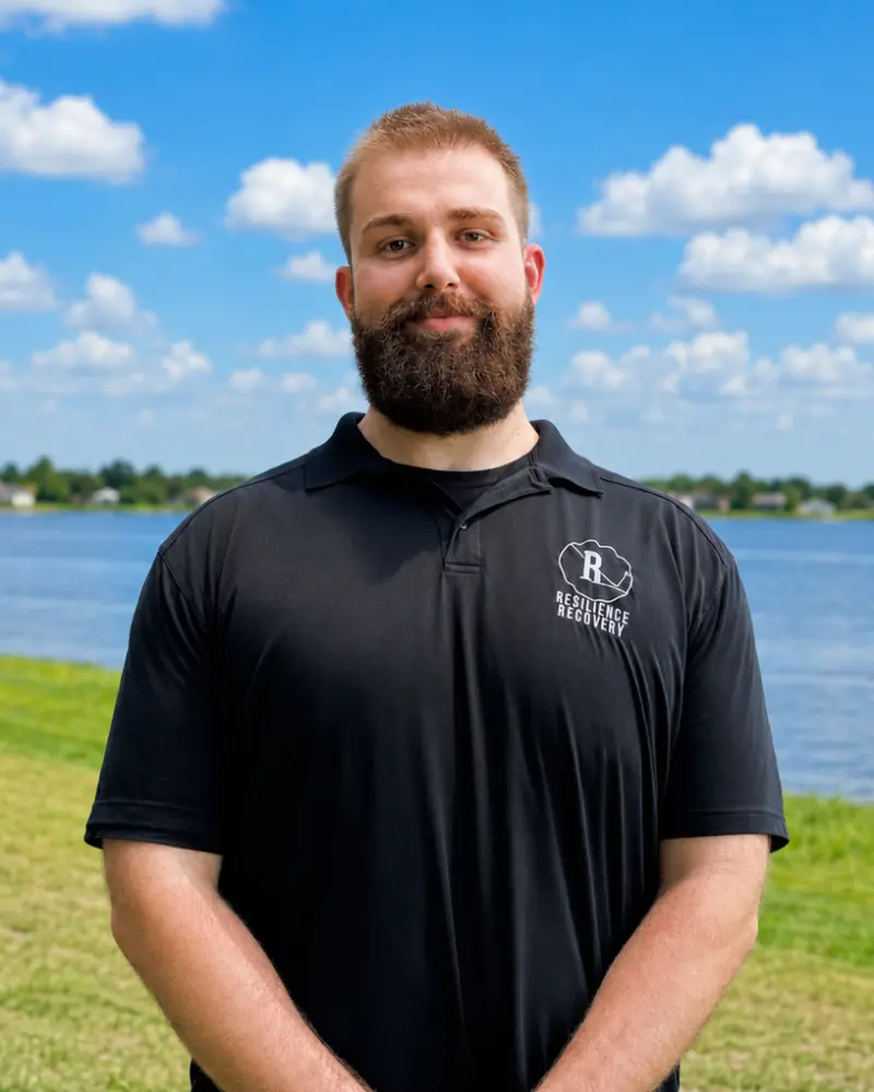A man with short hair and a full beard wearing a black polo shirt stands outdoors by a lake under a blue sky with scattered clouds.