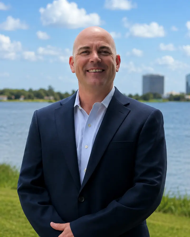 A smiling, bald man in a navy suit and white dress shirt standing outdoors by a body of water, with a blue sky and city buildings in the background.