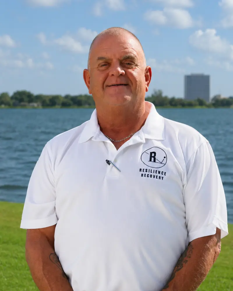 Smiling older man standing outdoors by a lake, wearing a white polo shirt with a “Resilience Recovery” logo, with blue sky and city buildings in the background.