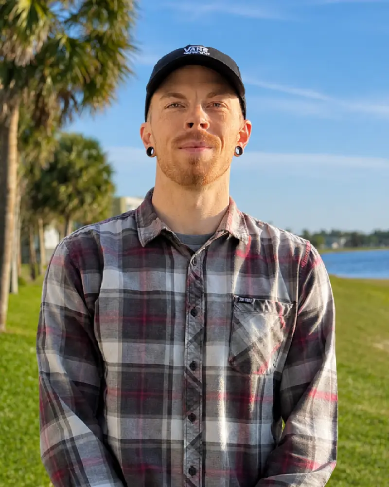A man outdoors wearing a dark baseball cap and plaid button-up shirt, standing in a grassy park with trees and a waterfront in the background.
