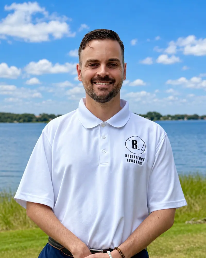 Smiling man standing outdoors by a lake, wearing a white polo shirt with a “Resilience Recovery” logo.