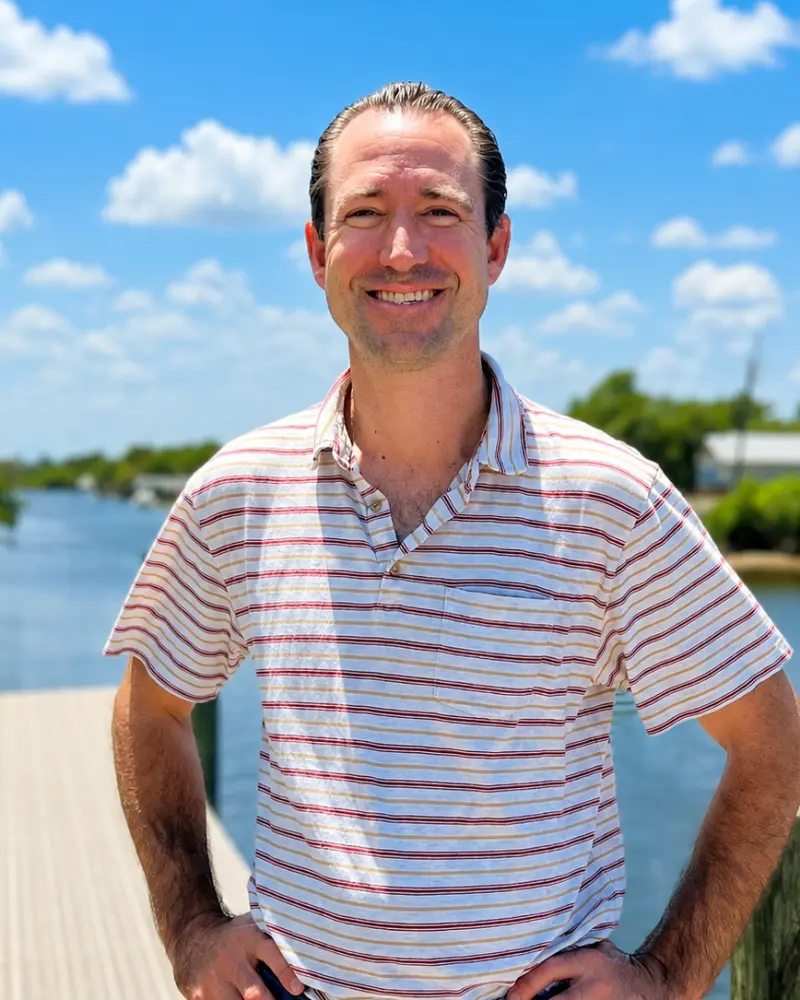 A smiling man in a striped polo shirt standing outdoors by a body of water under a blue sky with scattered clouds.