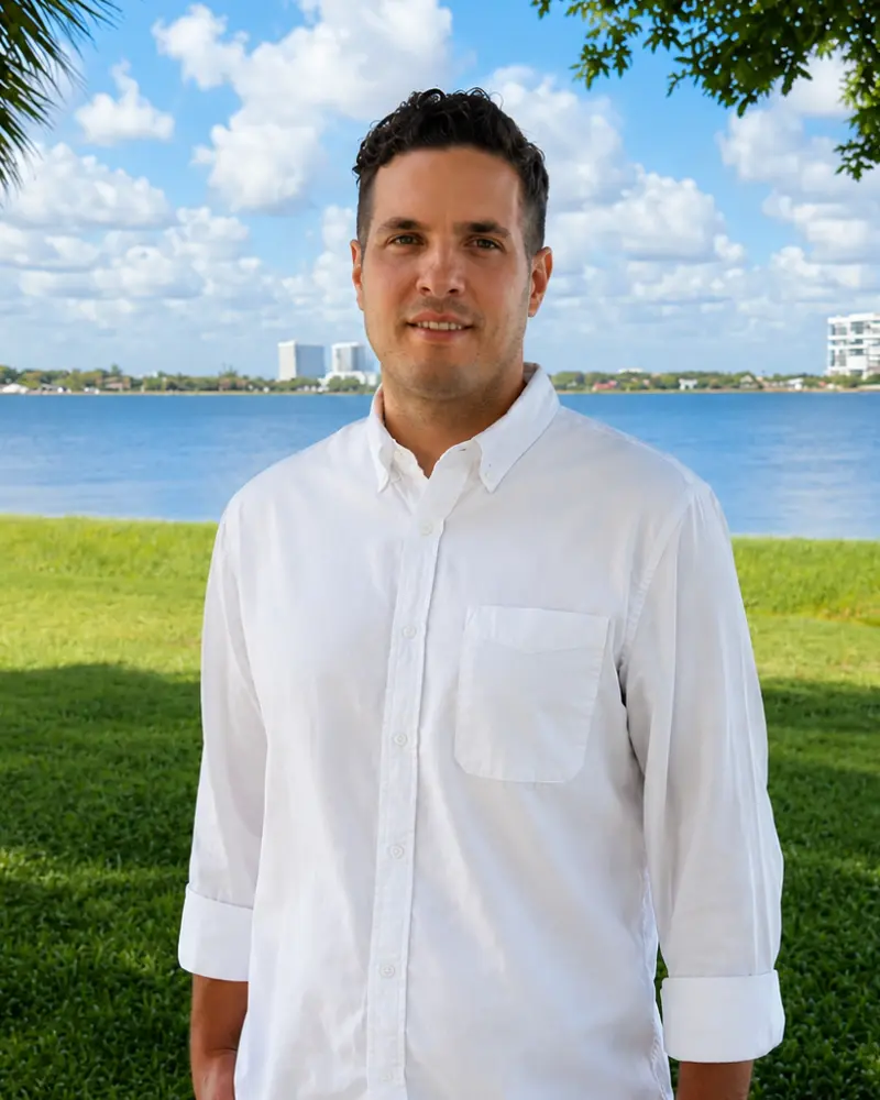 A man in a white button-up shirt standing outdoors near a grassy park by the water, with a blue sky and distant buildings in the background.