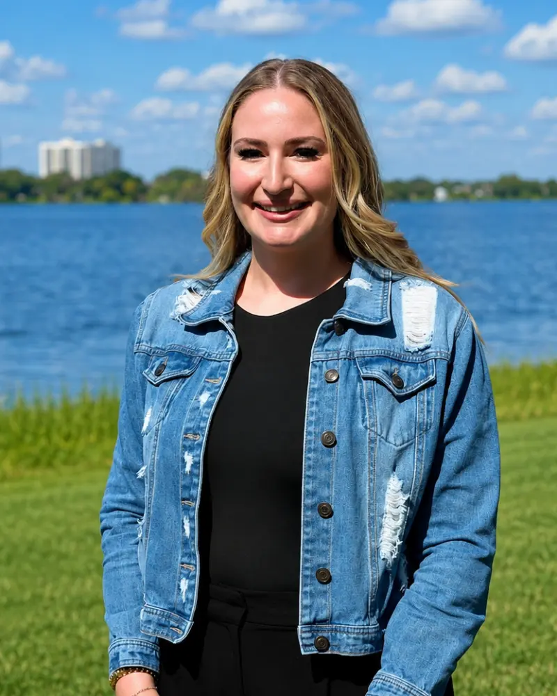 A smiling woman standing outdoors by a lake, wearing a blue denim jacket and black top.