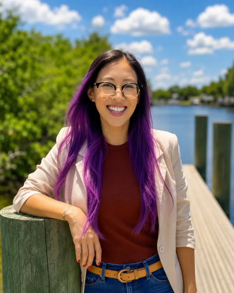 A smiling person with long purple hair and glasses standing by a wooden railing on a lakeside dock under a blue sky with clouds.