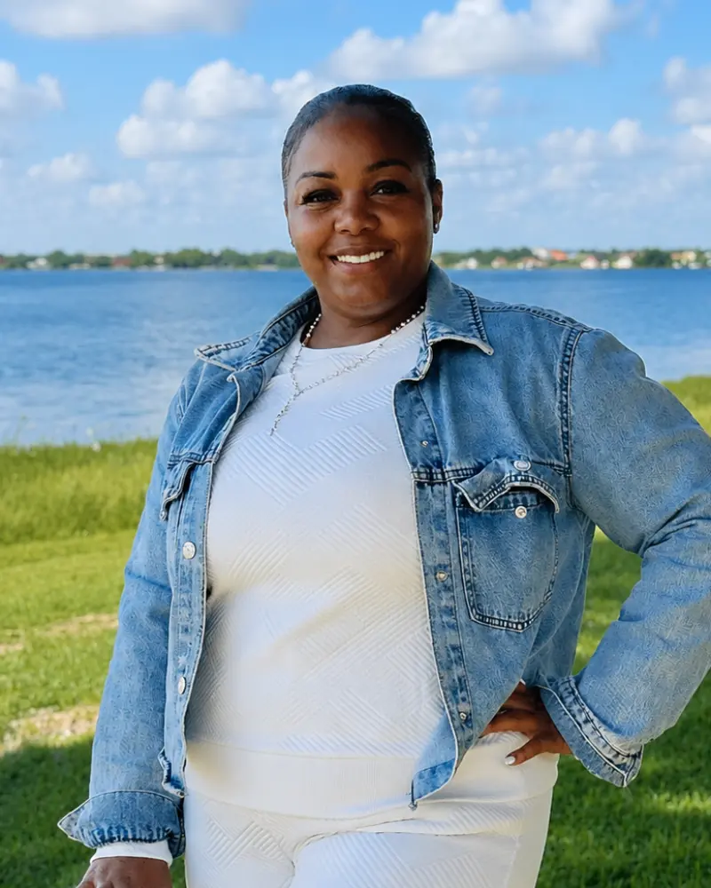 Smiling woman wearing a blue denim jacket and white outfit standing by a waterfront with green grass in the foreground and a blue sky with clouds behind her.