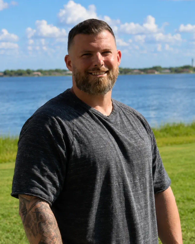 A smiling man with a short beard and short hair standing outdoors near a grassy shoreline, with a lake and blue sky in the background.